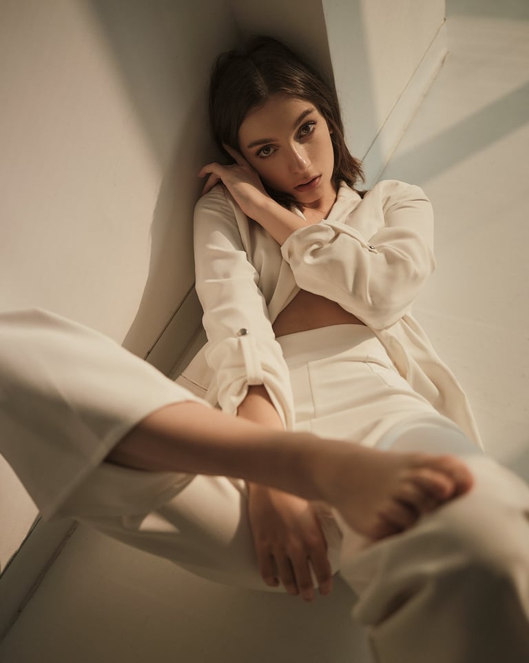Portrait of Masha laying indoors wearing a white suit, leaning against a wall in soft natural light.