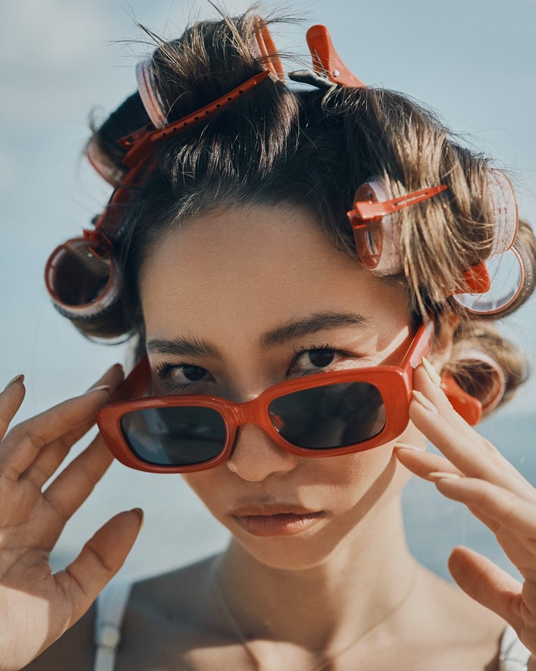 Close-up portrait of Yuki wearing red sunglasses with hair set in rollers, photographed outdoors in daylight.