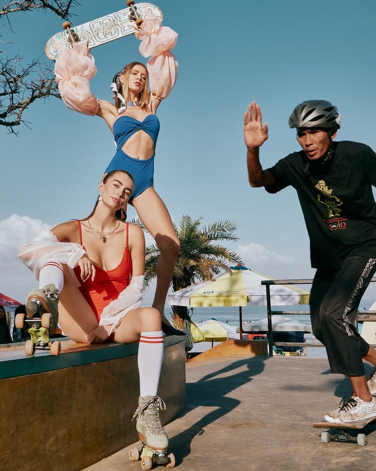 Two models in red and blue swimwear pose with roller blades and a skateboard at a Bali skate park as a man passes.