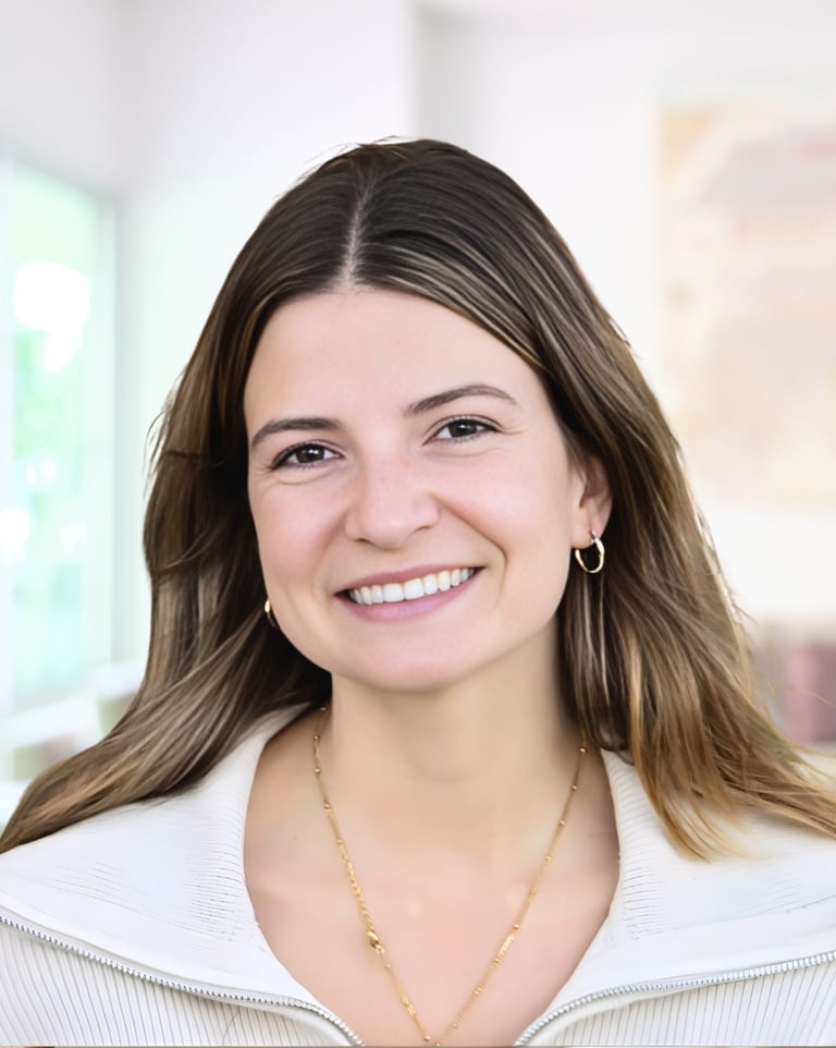Professional headshot of a smiling young woman with brown hair, gold jewelry, and a white blazer.