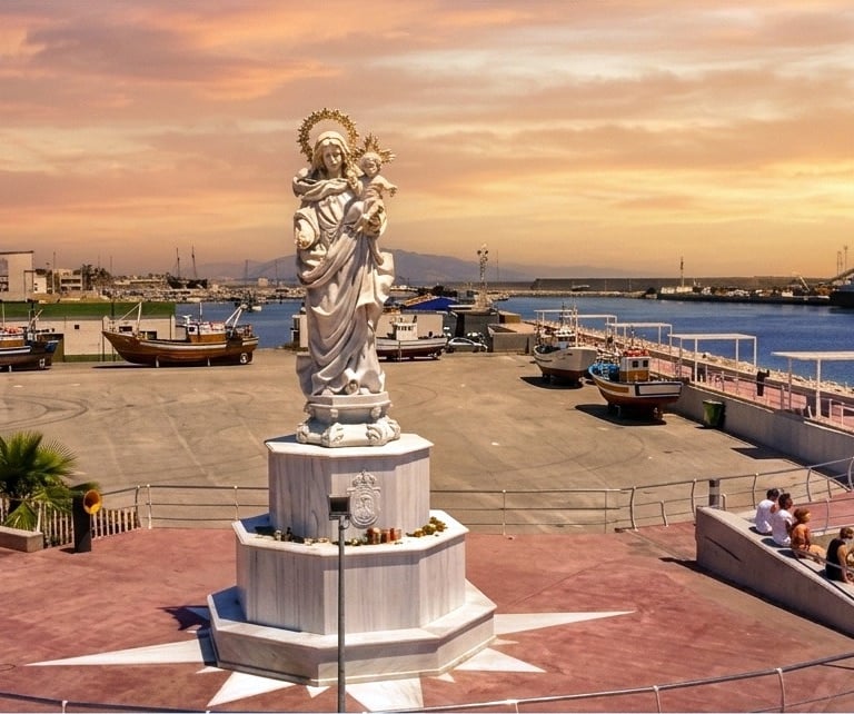 A white marble statue of the Virgin Mary overlooking a harbor with fishing boats at sunset.