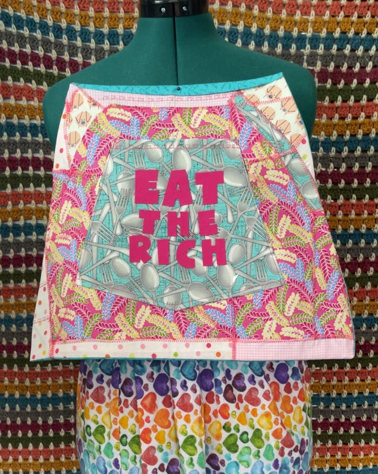 An Apron art piece that says EAT THE RICH in pink letters over utensils fabric accented with pig fab