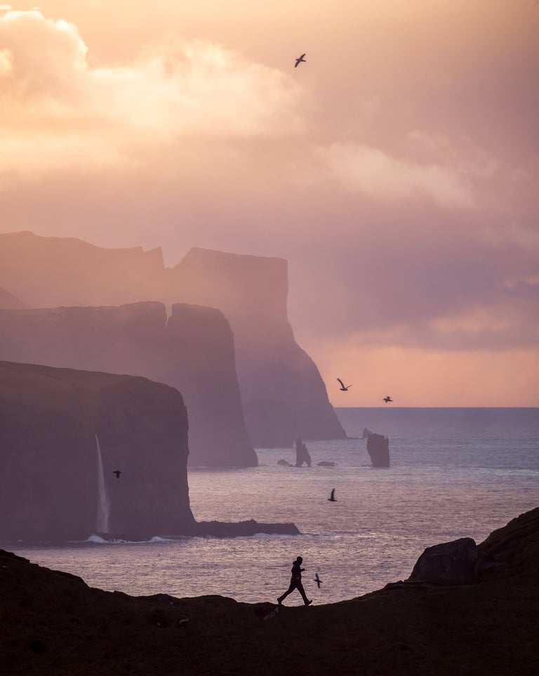 Man running along Kallur lighthouse cliffs with dramatic drops and ocean below, wild Faroe Islands landscape