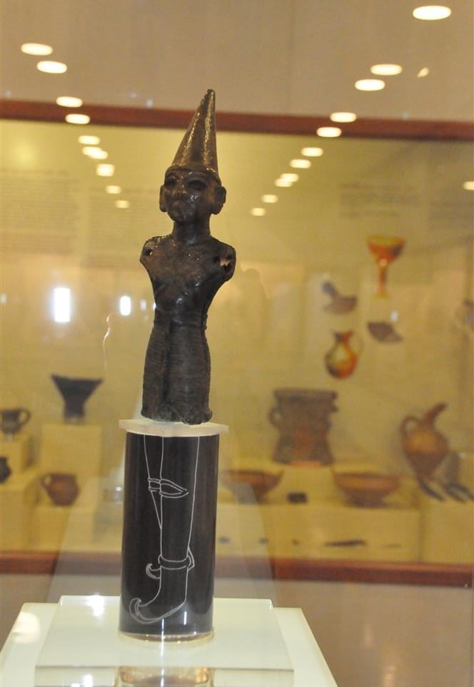 Ancient bronze Hittite deity statuette wearing a conical hat in a museum display case.