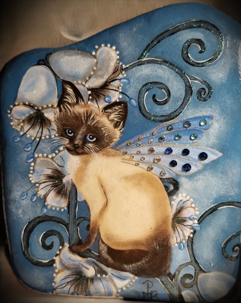 A rock painting of a Siamese Fairy Kitty on blue bell flowers.