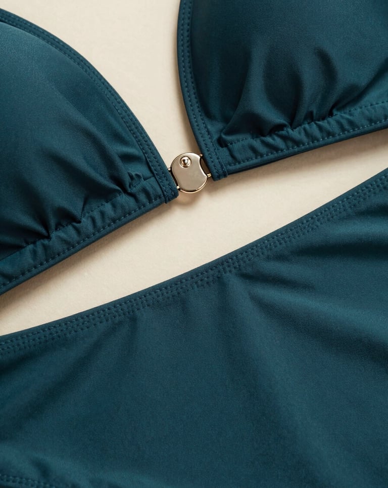 A high-fashion macro shot focusing on the intricate stitching and hardware of a luxury swimsuit. The deep charcoal teal fabric is contrasted by a soft warm cream light, showcasing the premium craftsmanship.