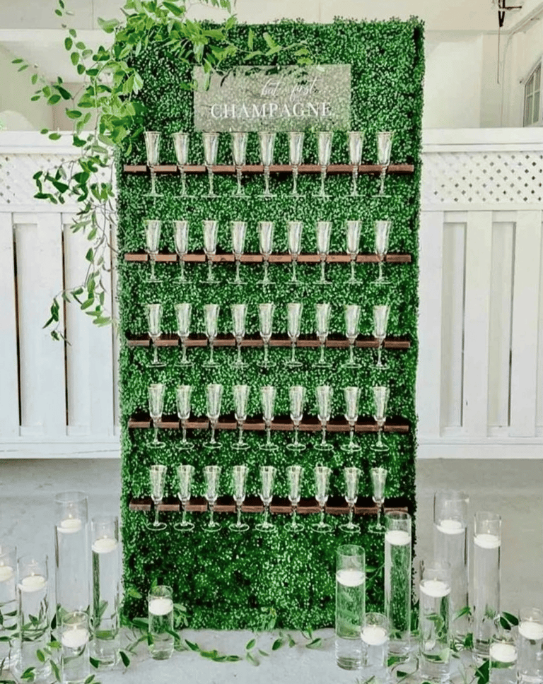 A wall adorned with lush greenery with a "But first Champagne" sign for a wedding.