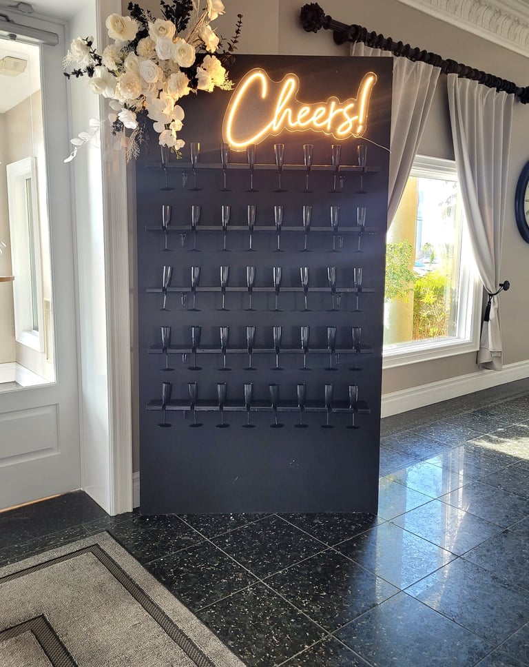 Matte black champagne wall with black champagne glasses, white floral arrangement and a cheers neon