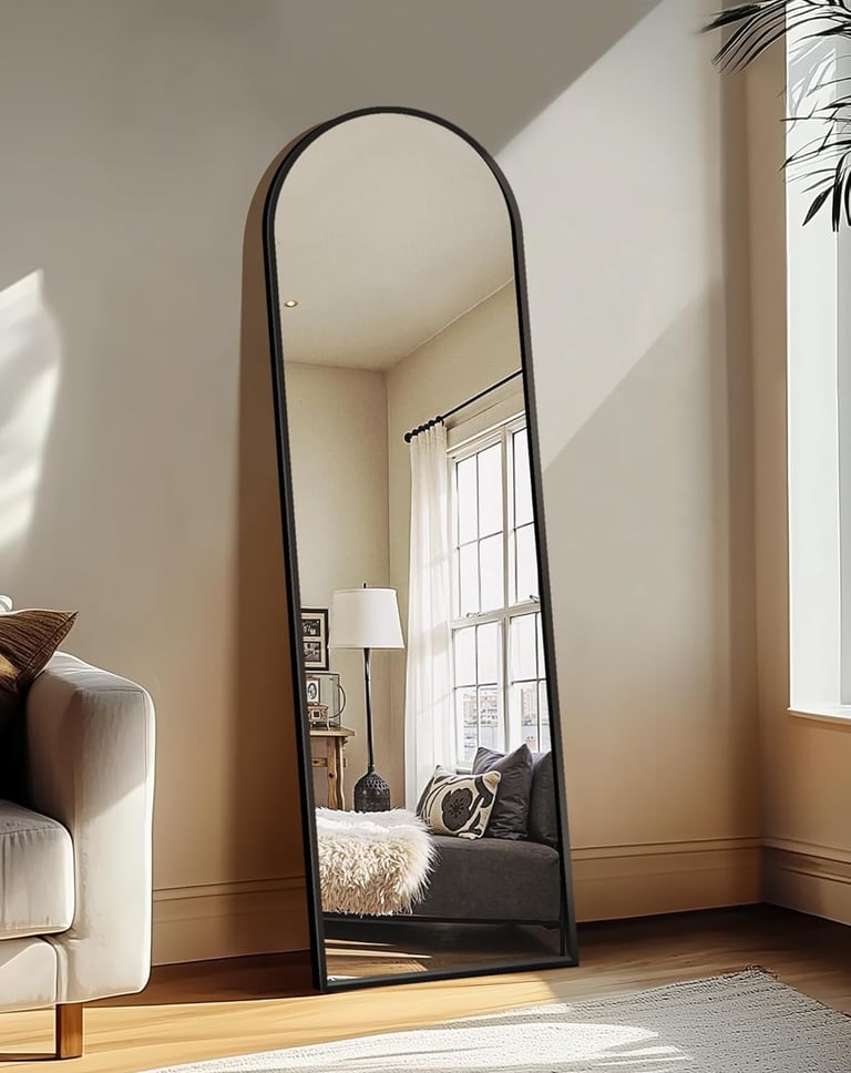 Full-length arched floor mirror with a black metal frame leaning against a wall in a bright modern living room.