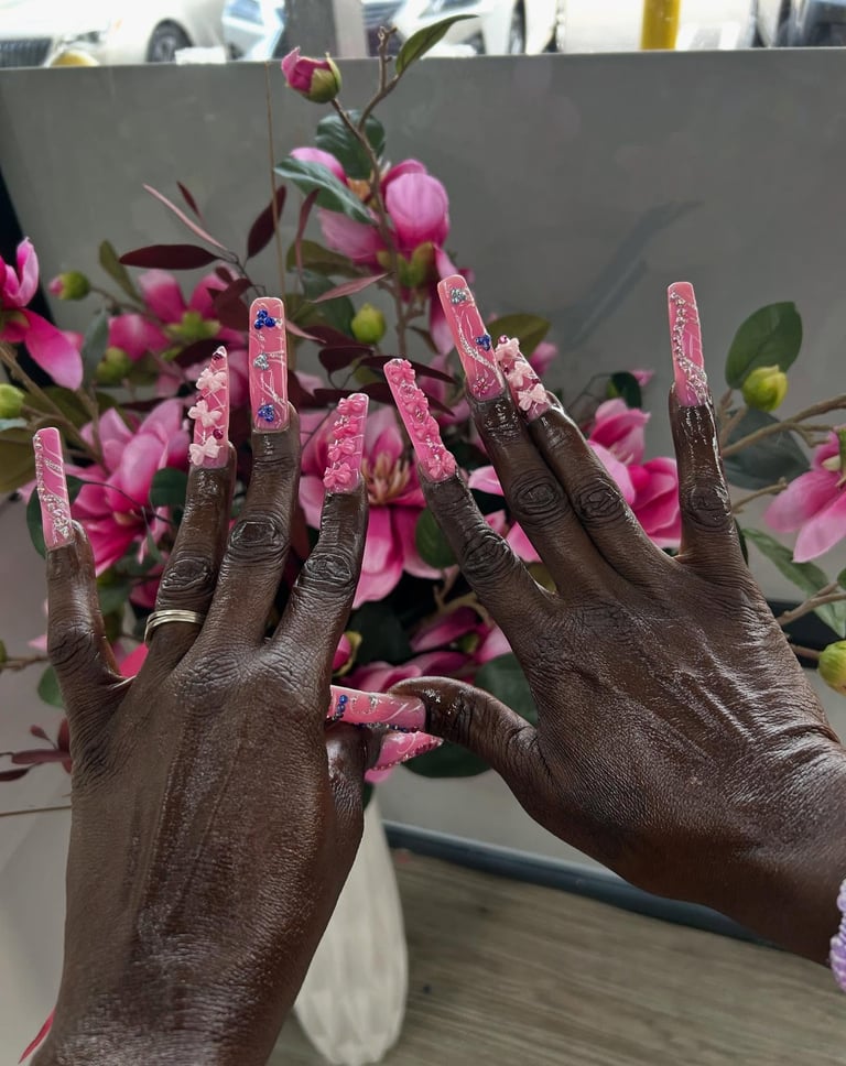 Extra long pink acrylic nails with 3D floral art and glitter accents in front of flowers.