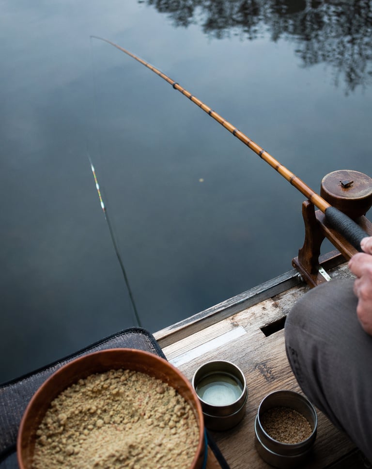 A fisherman holds a bamboo fishing rod over calm lake water with bait bowls nearby on a wooden dock.