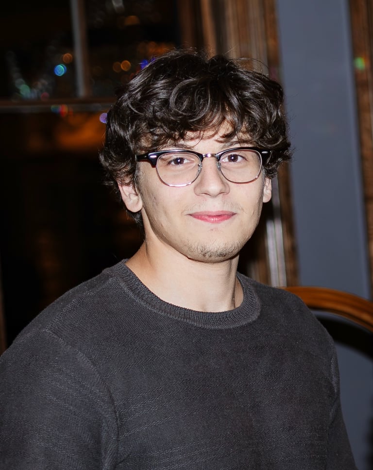 Young man with curly brown hair and glasses wearing a dark sweater in a portrait.