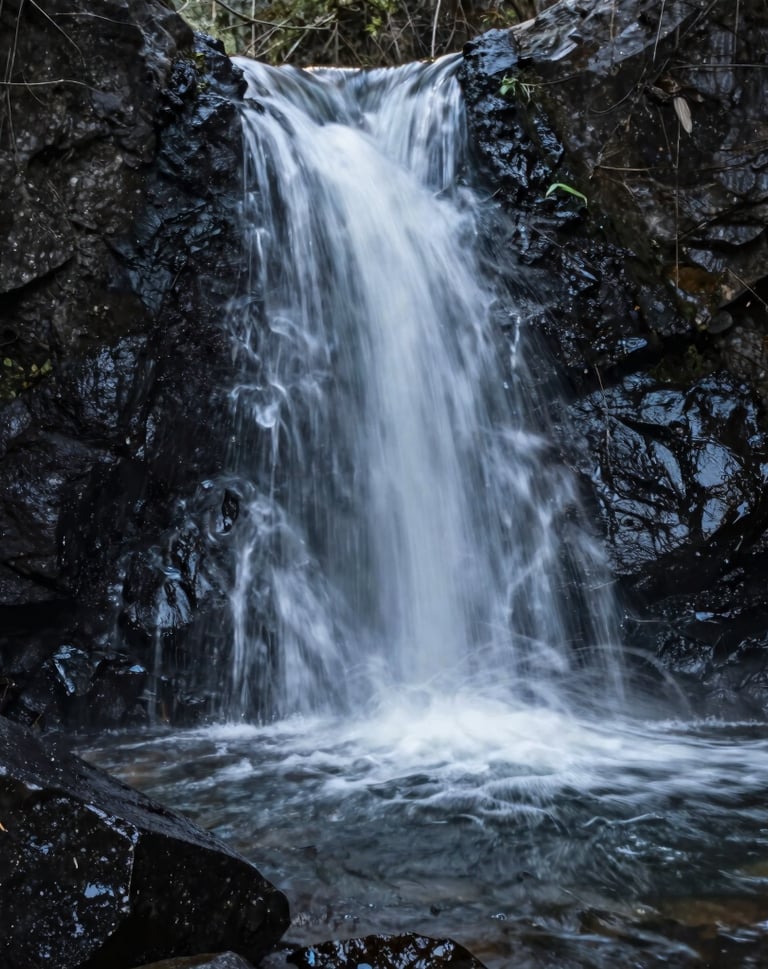 A vertical shot of a minimalist waterfall in a North American forest. The water is a soft motion blur against dark near black wet stones. Clean, calming aesthetic with muted organic tones.
