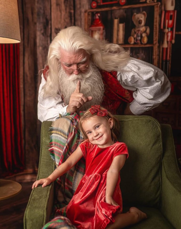 Santa Claus making a quiet gesture behind a smiling young girl sitting in a green armchair.