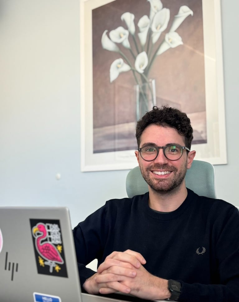 Smiling man with glasses sitting at a desk with a laptop in a modern office setting.