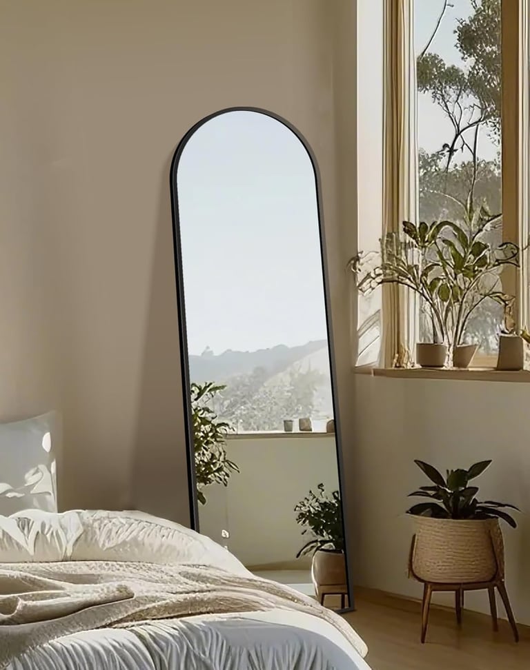 A full-length arched floor mirror with a black frame leaning against a wall in a bright, modern bedroom.