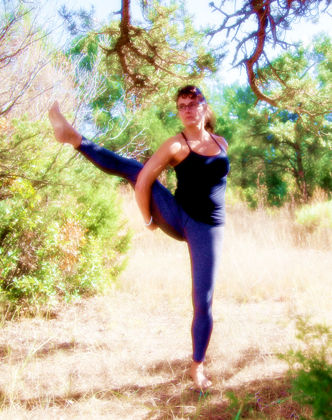 A woman practices a standing balance yoga pose in a sunny forest clearing for outdoor wellness.