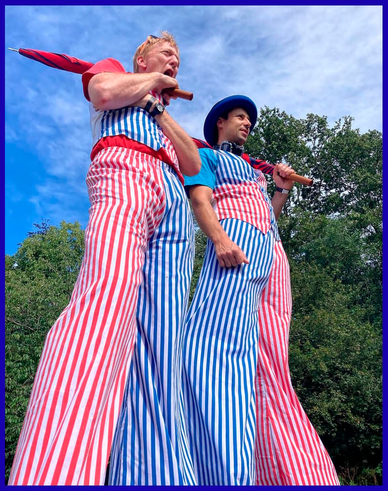Two circus performers on stilts wearing red and blue striped trousers and holding umbrellas.