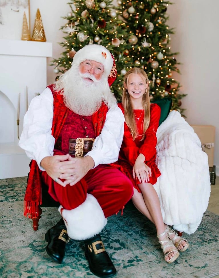 Smiling girl sitting with Santa Claus in front of a decorated Christmas tree with lights.