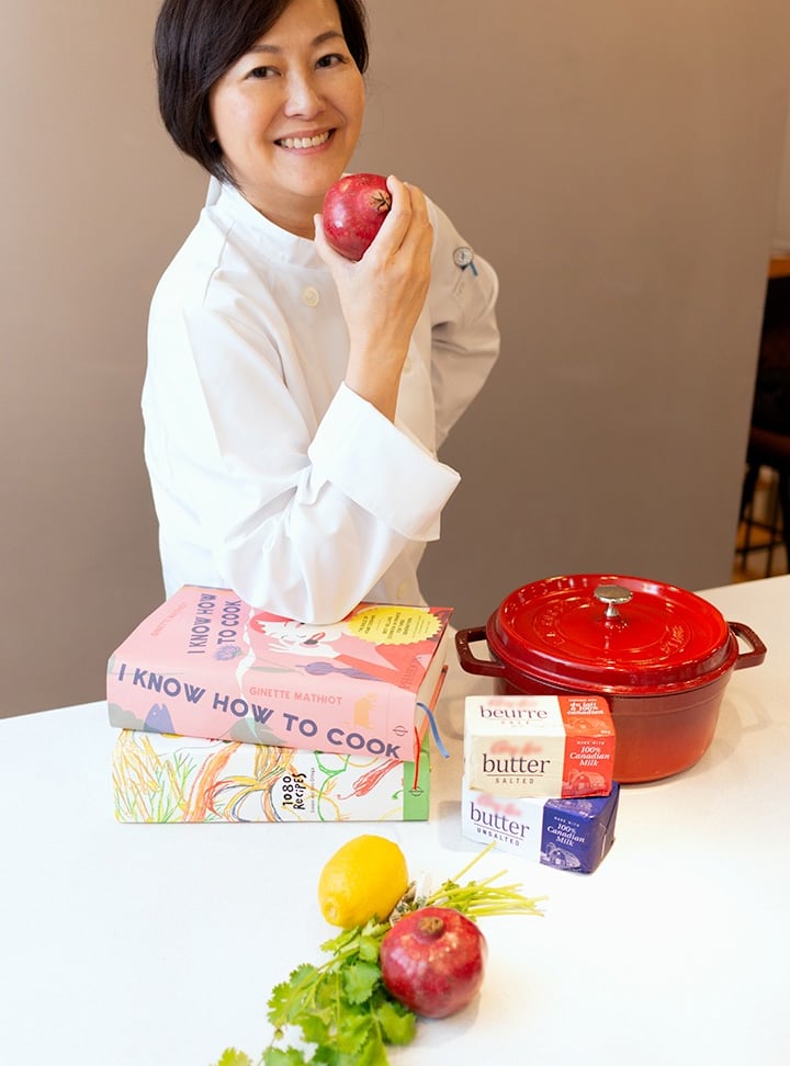 a woman in a chef's uniform holding a red pomegranate smiling at the camera