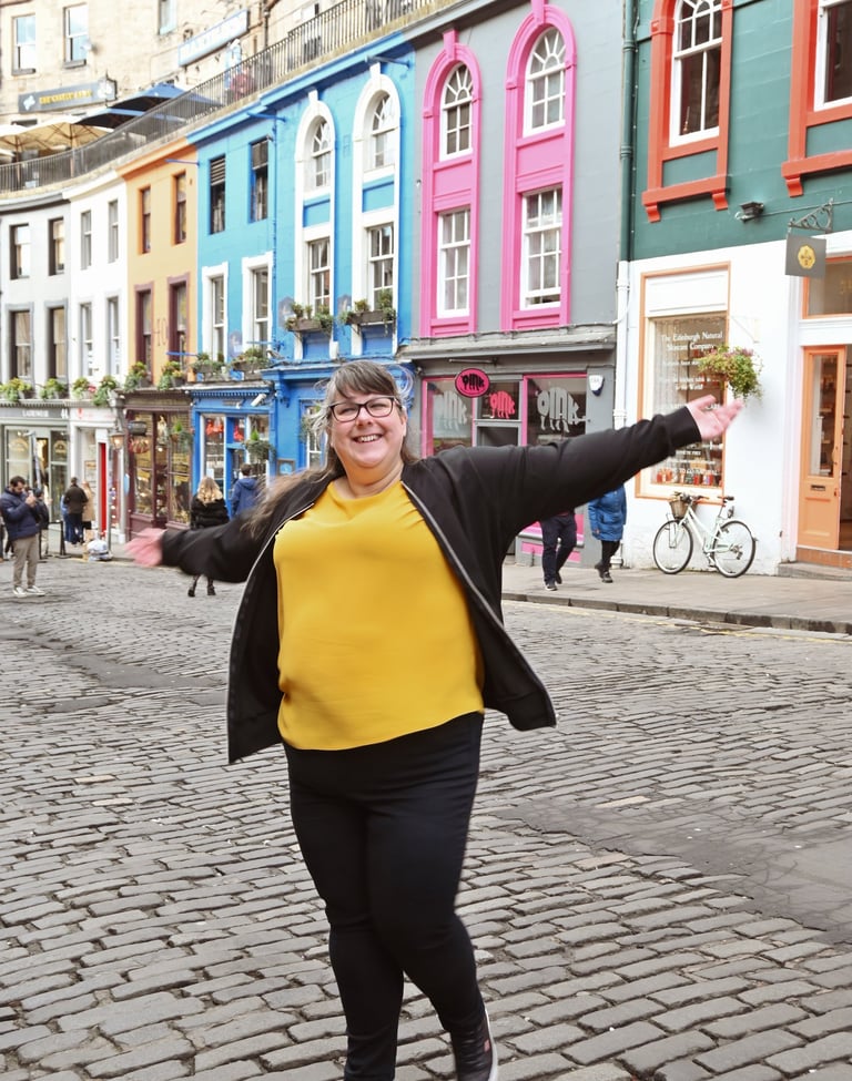 Laura is dancing with her arms out on a cobbled a street with brightly coloured buildings behind her