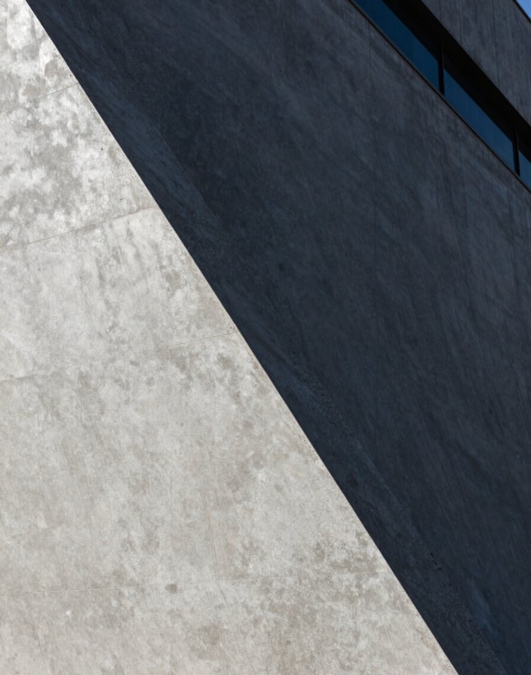 A detailed close-up shot of architectural textures. A smooth light silver grey concrete surface meets a dark charcoal grey metallic finish. Harsh sunlight creates a dramatic diagonal shadow across the frame, emphasizing the high-contrast aesthetic of a modern South American / Brazilian building.