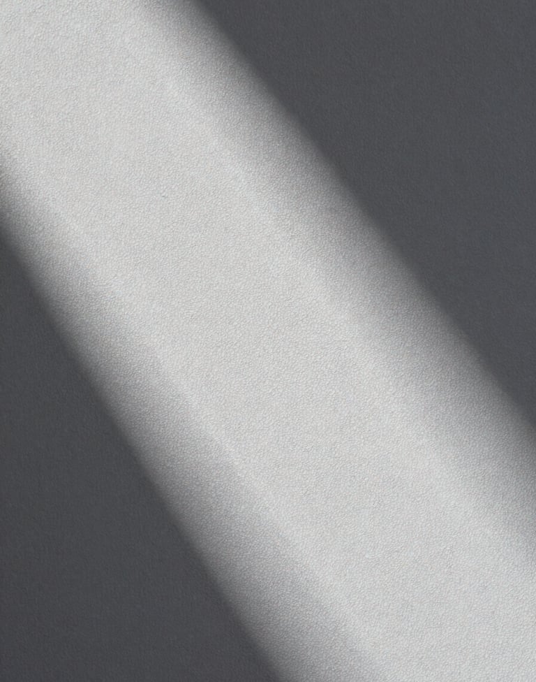 A close-up photographic study of light hitting a white textured wall. The composition is abstract, focusing on the subtle gradients of gray and the tactile quality of the surface. Minimalist and architectural, North American / European style.