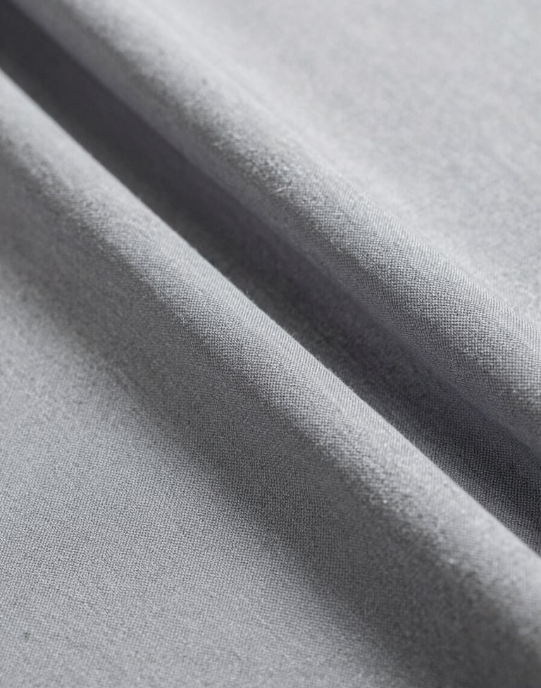 A close-up photograph of high-quality textile folds in shades of light gray and medium gray. The lighting is soft and diffused, capturing the delicate weave and sophisticated texture of the material in a clean, artistic composition.