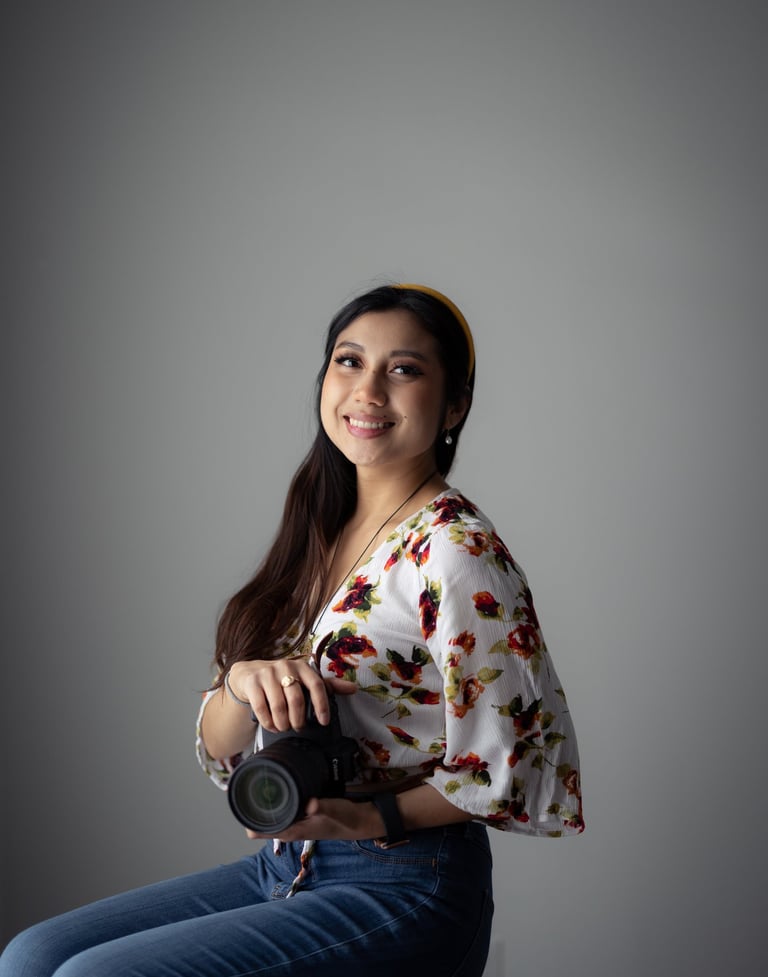 Professional shot of me holding Canon camera, sitting on a stool. Wearing floral top and headband.