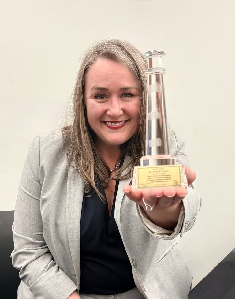 A smiling woman in a gray blazer holding a glass lighthouse award for professional excellence.