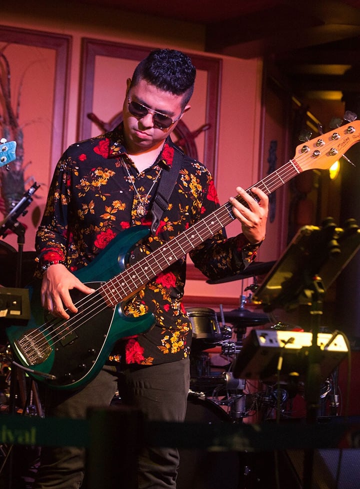 Bassist Ian Yoshio wearing a black shirt with red flowers, playing a navy blue Sterling bass guitar.