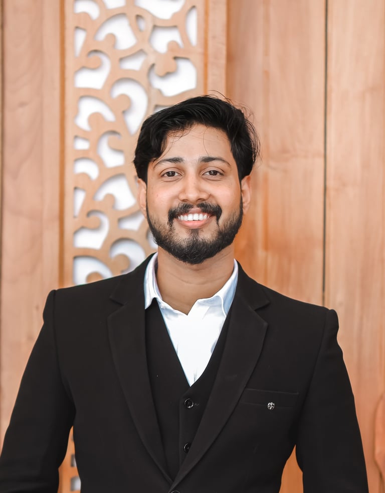 Smiling young man in a black formal suit and white shirt standing against a carved wooden background.