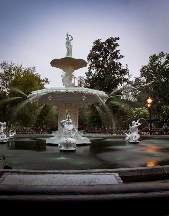 The historic Forsyth Park Fountain in Savannah, Georgia, featuring white ornate statues and spraying water.