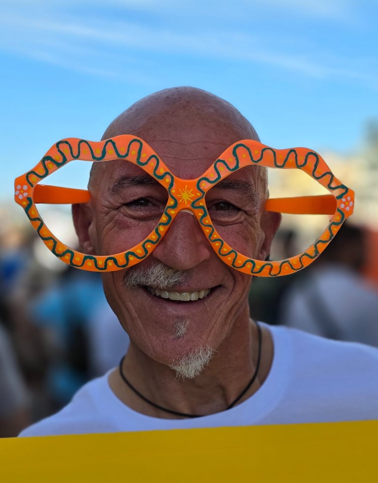 Older man smiles, wearing big colorful lips-shaped glasses outdoors, blue sky above and a blurred crowd behind him.