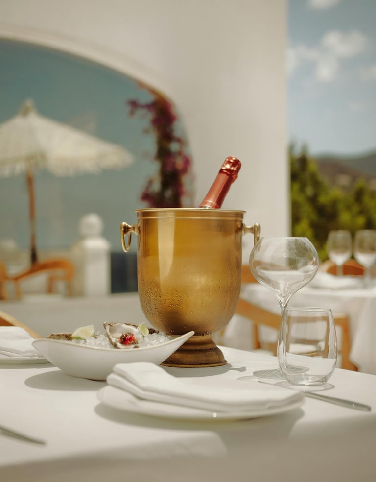 Outdoor dining table with champagne in an ice bucket, oysters on a plate, and empty glasses.