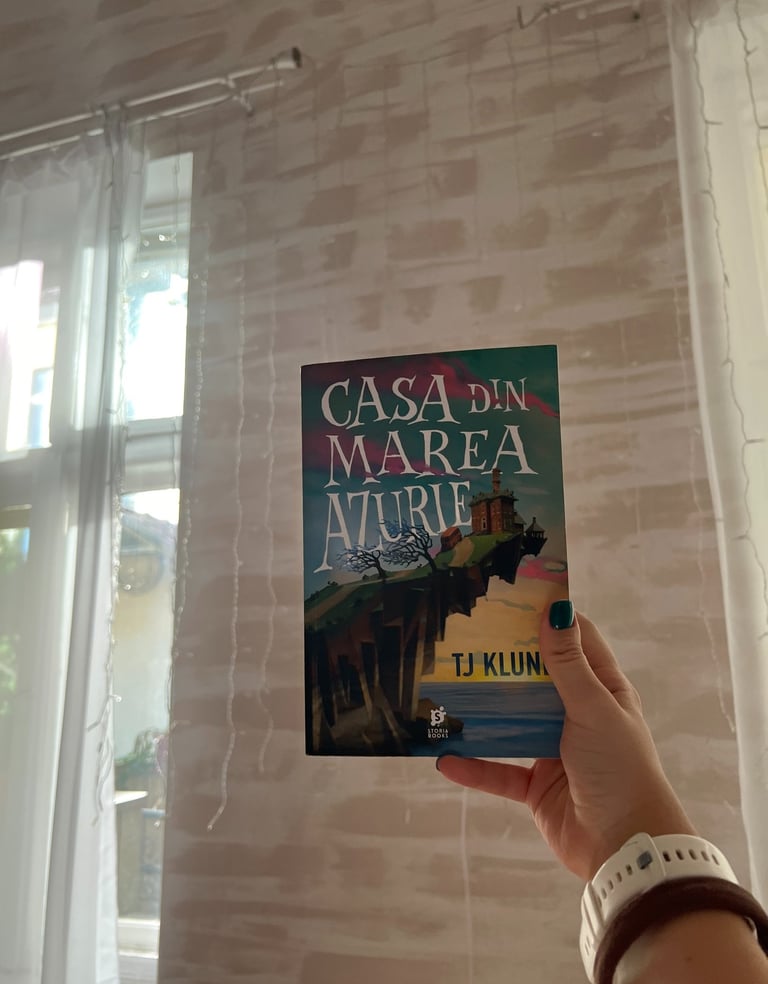 Mână ținând cartea 'Casa din Marea Azurie' de TJ Klune pe un fundal de luminițe de basm.