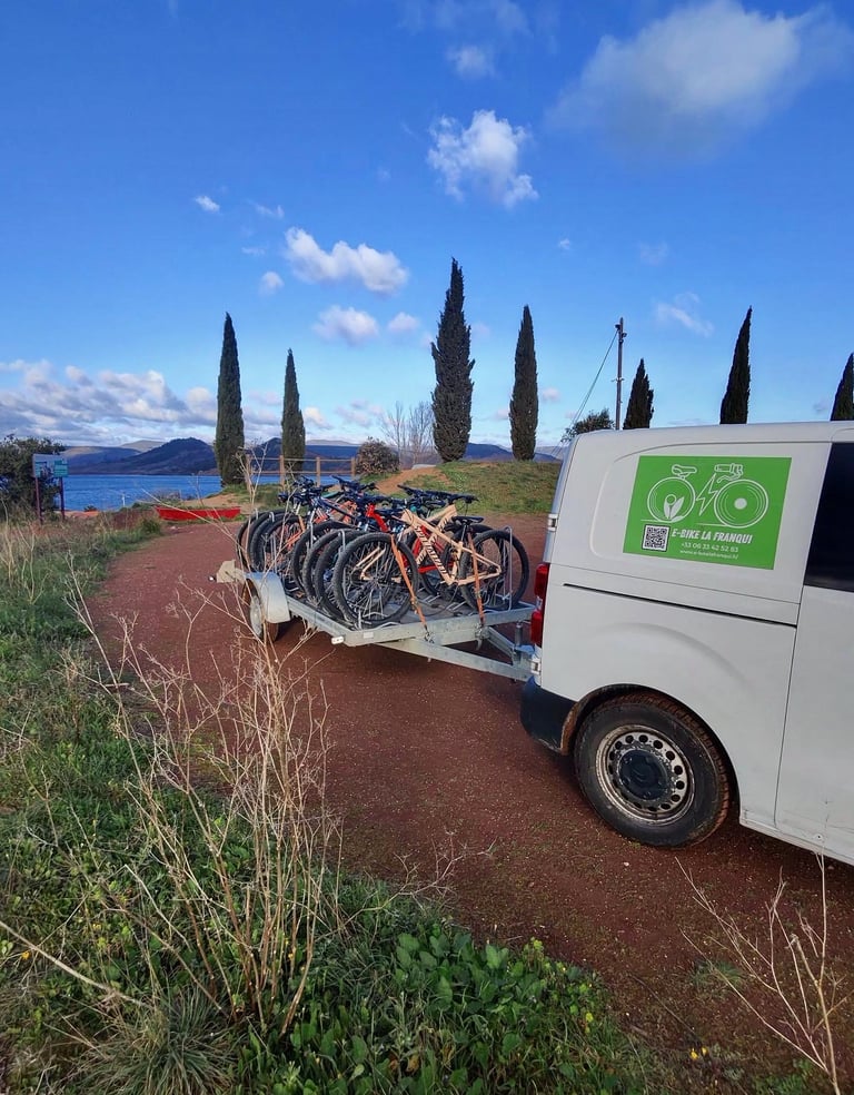 White E-Bike van pulling a trailer loaded with mountain bikes on a scenic path near a lake.