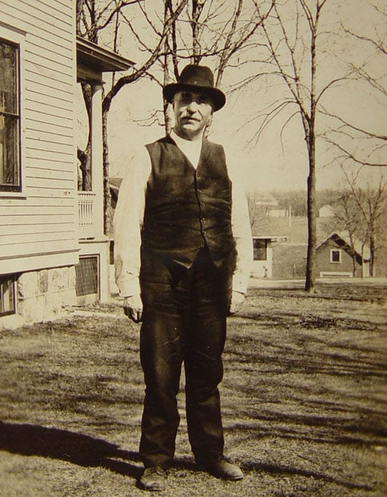 a gentleman in a vest and hat standing outside a house, black and white