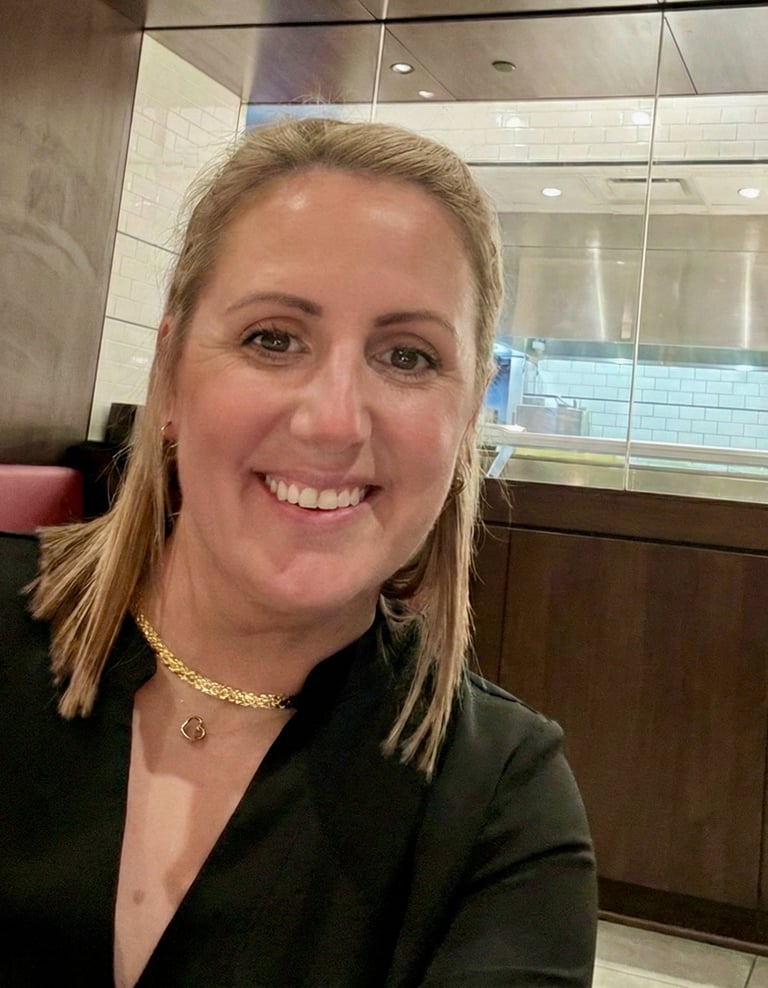 Smiling woman with blonde hair wearing a gold necklace in a modern restaurant setting.