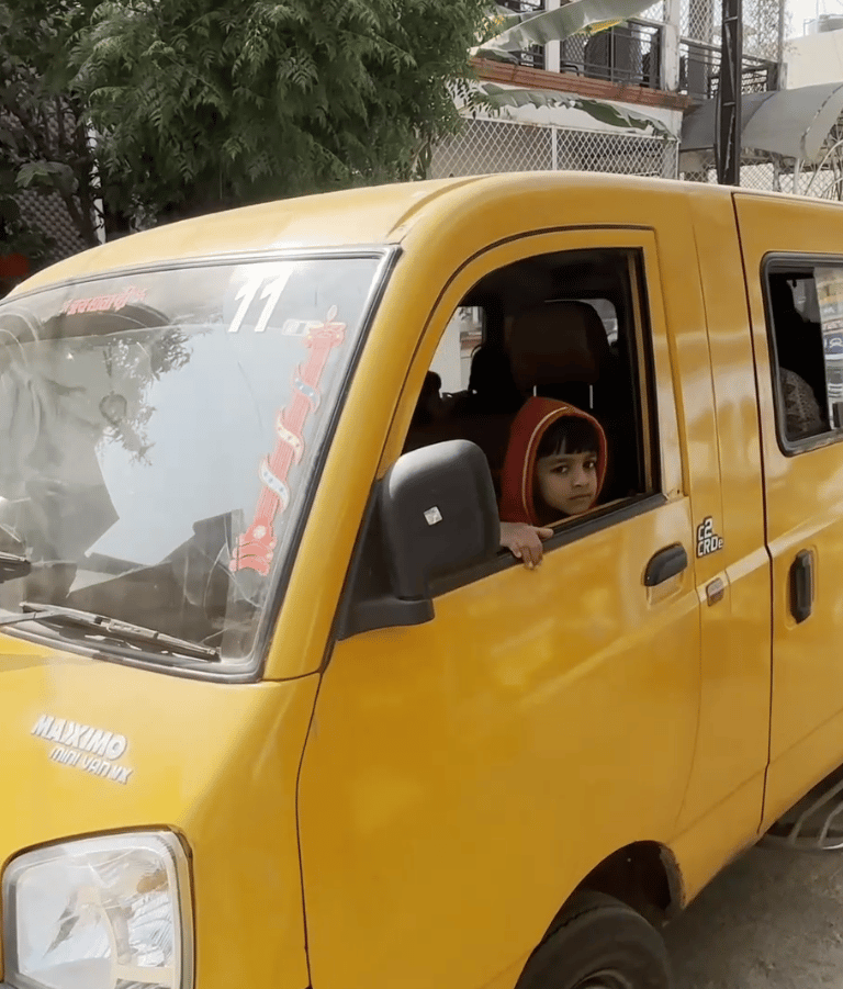 A young child in a red hoodie looking out of a yellow Mahindra Maxximo mini van on a city street.