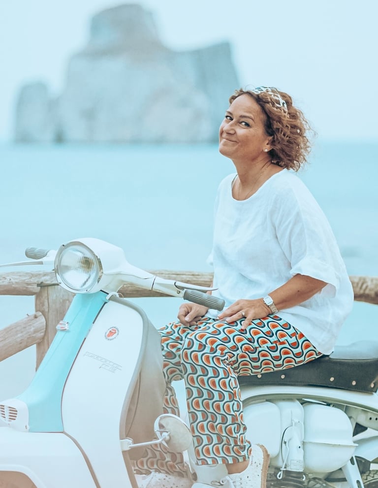 Alessandra Polo on a Vespa with view of Pan di Zucchero sea stack in Sardinia.