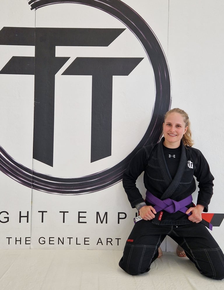 A female martial artist in a black BJJ gi and purple belt kneeling in front of a gym logo.