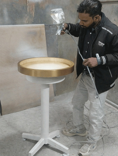 Professional painter applying a gold finish to a circular metal table using a spray gun.