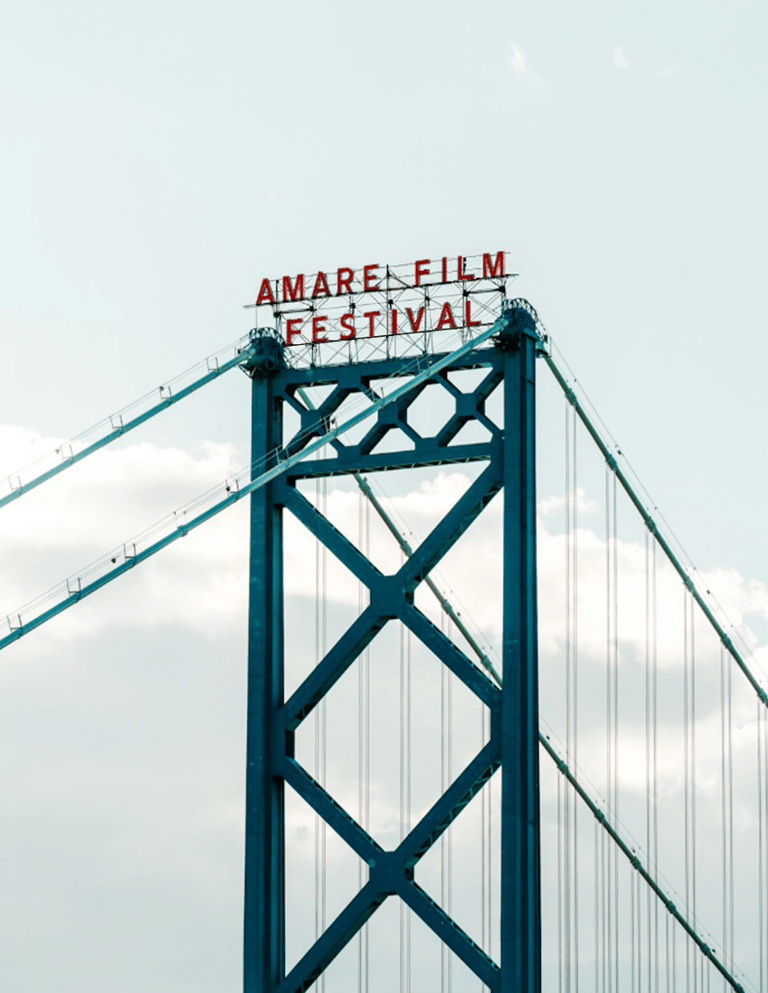 Red Amare Film Festival sign atop a blue steel suspension bridge tower against a cloudy sky.