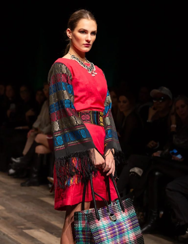 A model walks a fashion runway wearing a vibrant red dress with embroidered fringe sleeves and a woven tote bag.