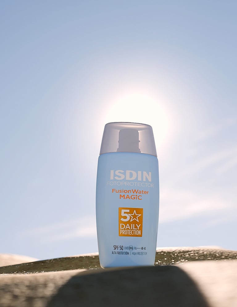 Bote de crema de sol de ISDIN en la arena y el sol detrás.