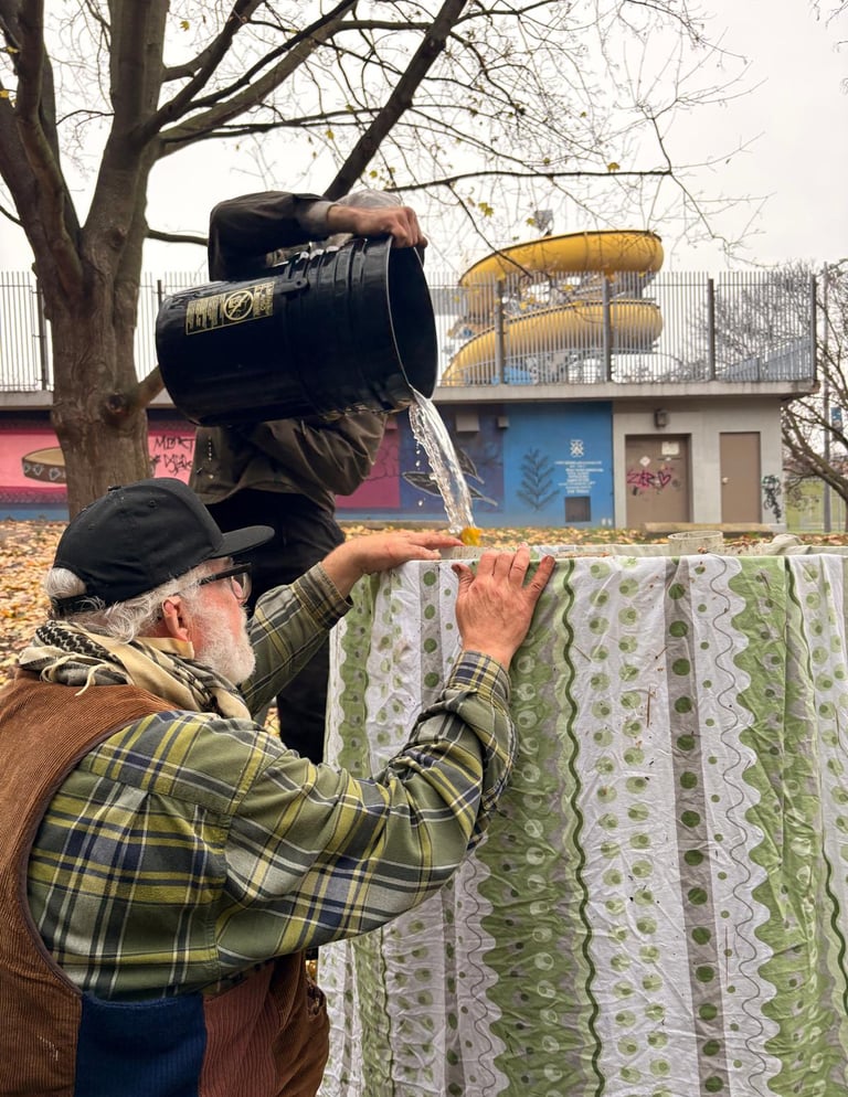 A man pours water from a black bucket over a fabric-covered structure in an urban park.