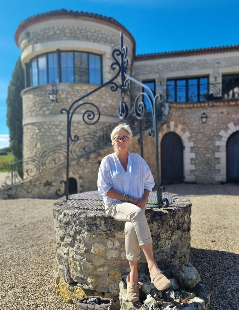 A woman sits on an old stone well in front of a historic European stone building with a round tower.