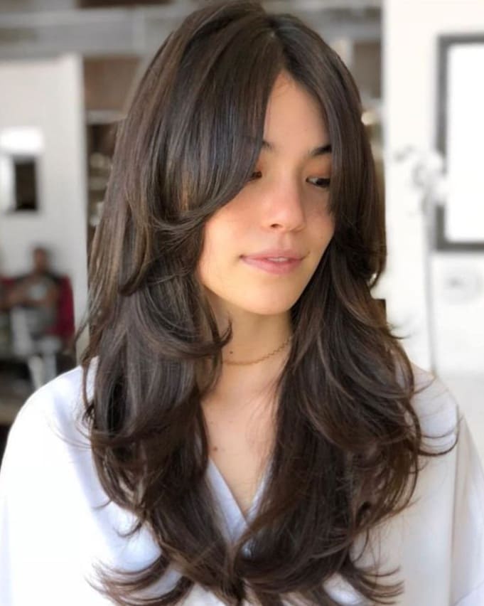 Mujer con corte de cabello largo en capas y flequillo tipo curtain bangs en tono castaño oscuro.