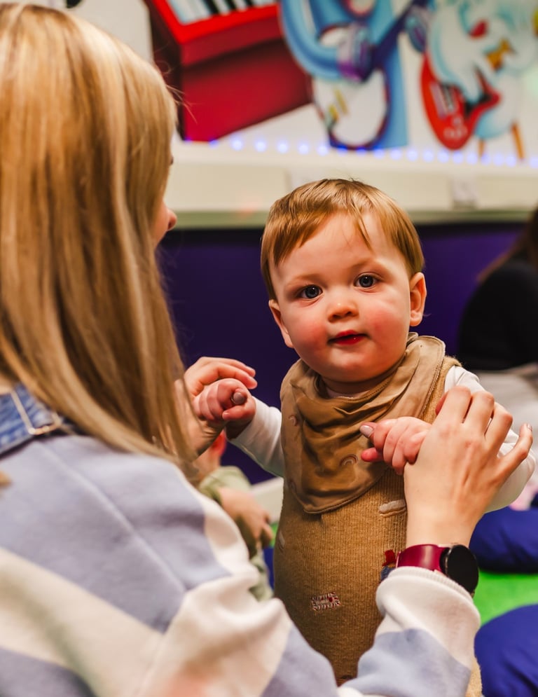 A young boy staring into the camera, holding his mums hands to stand up in moo class.